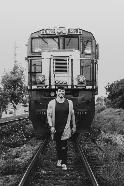 Young man smiling in front of a train on railway tracks in Pinhais, Brazil.