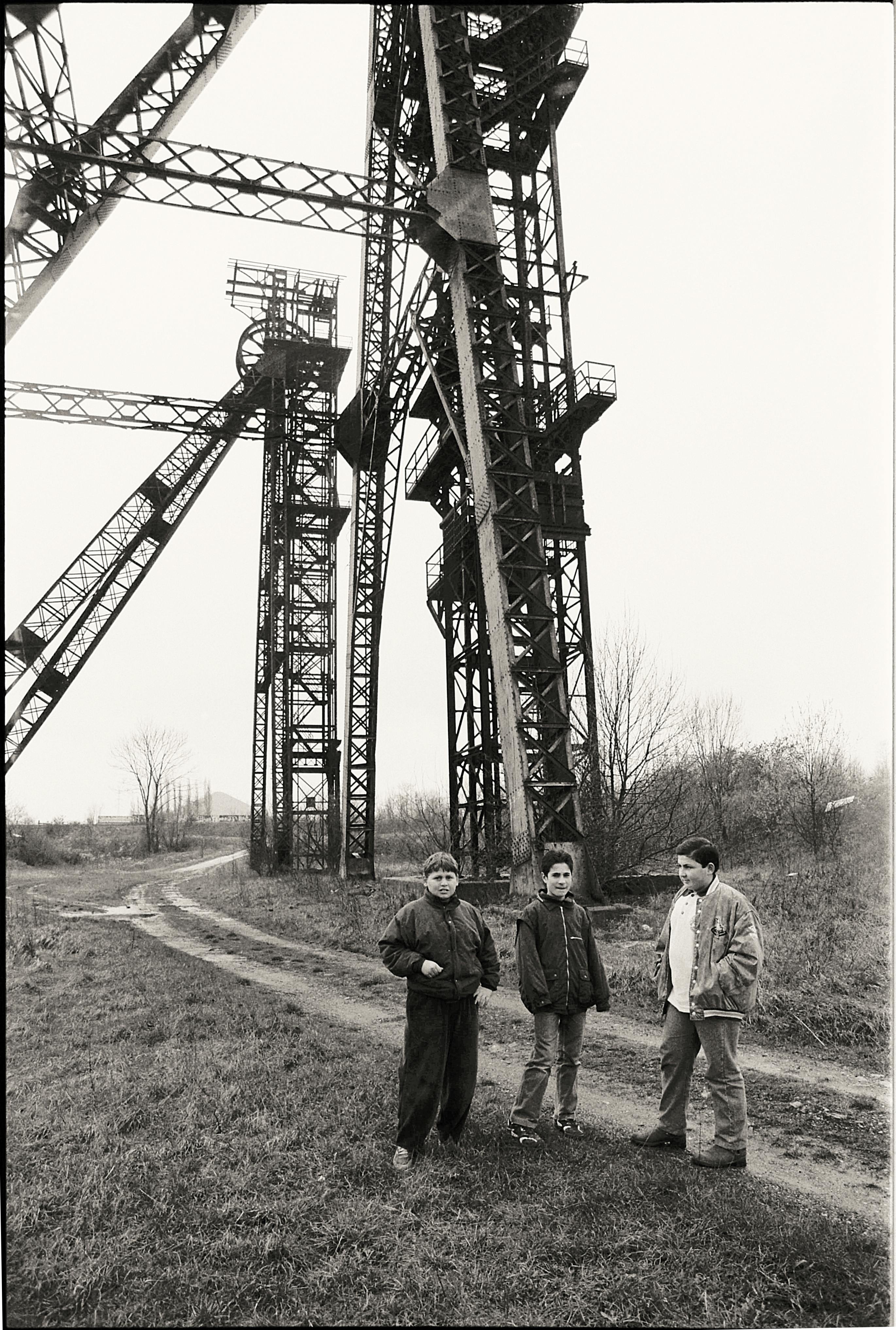 Black and white image of three boys standing near industrial metal towers in a field.