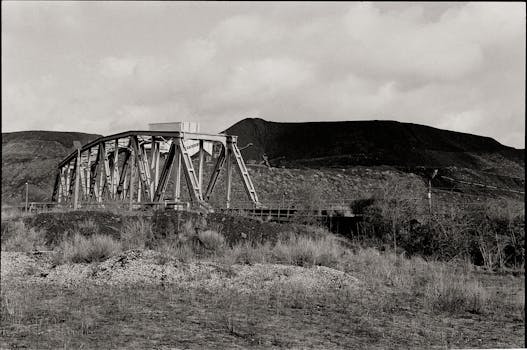 Monochrome image of a metal bridge with hills and dry grass.