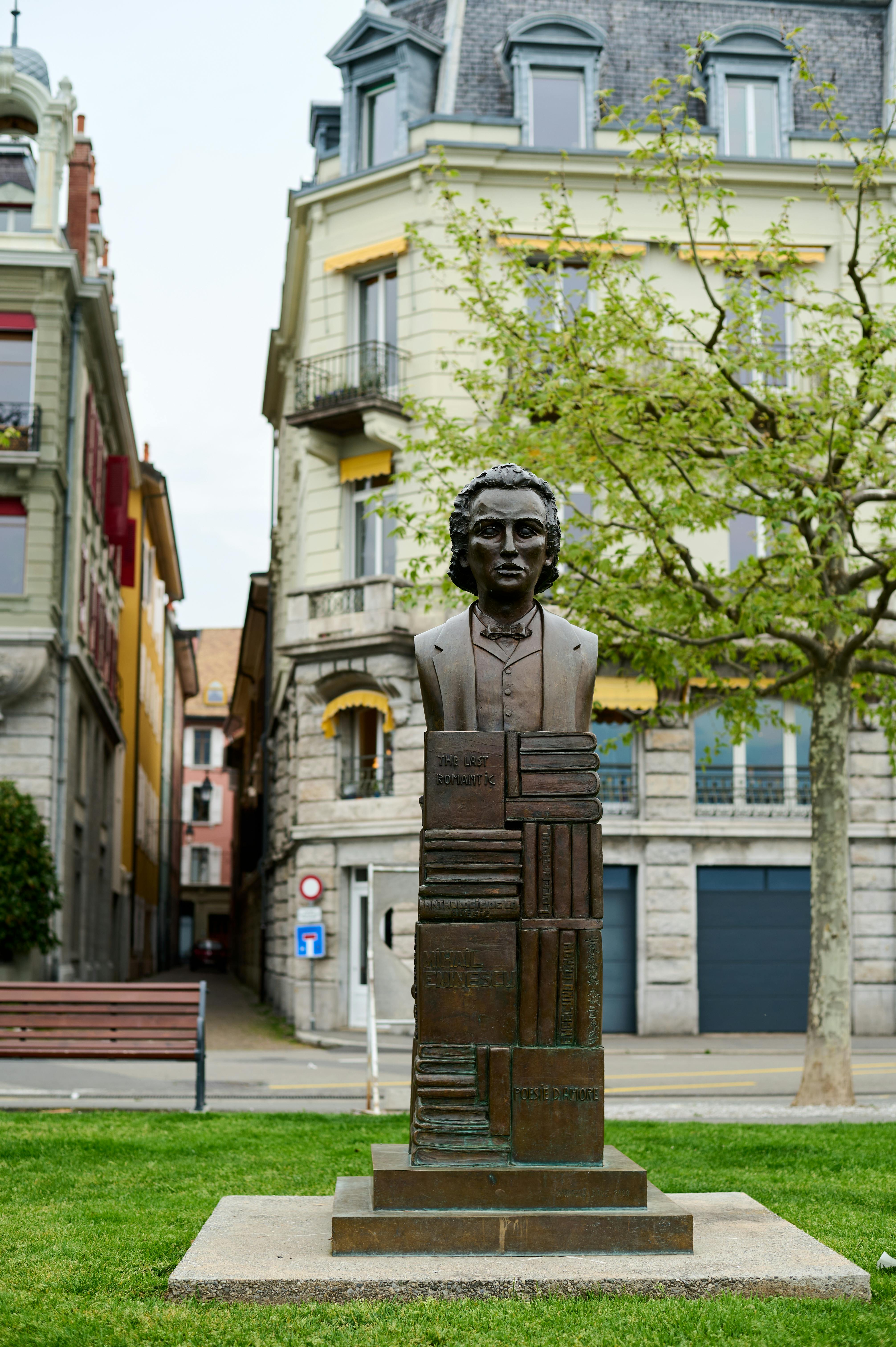 A striking statue in a city park with historic buildings and lush greenery.
