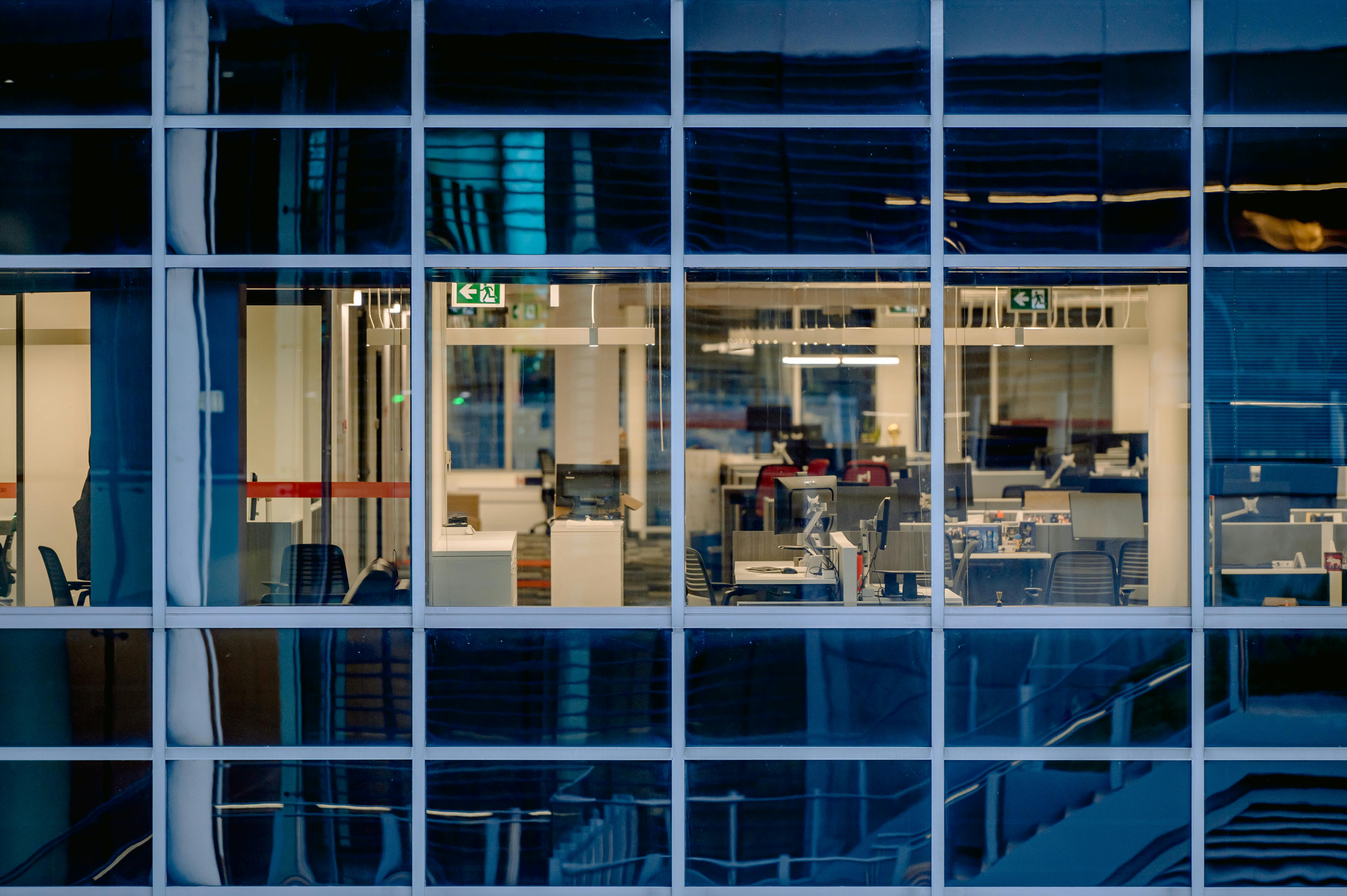 A modern office interior with empty desks viewed through blue-tinted windows at dusk.