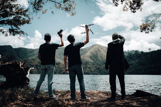 Three men holding drones by a mountain lake, silhouette against a scenic backdrop.