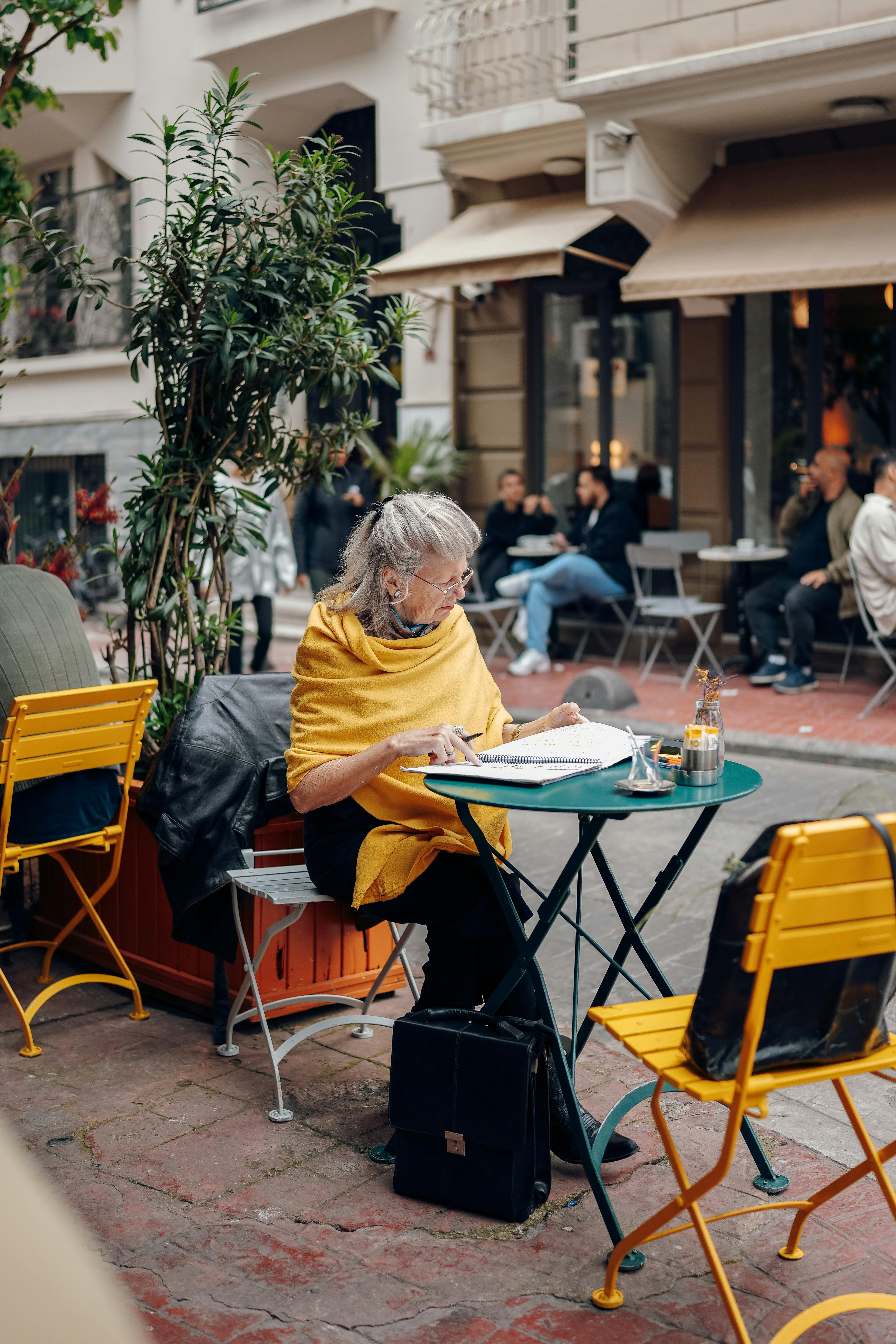 Senior woman enjoys reading while seated at an outdoor café in a bustling urban setting.