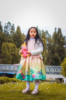 A young girl with long black hair holding a colorful toy stands outdoors in a scenic area.
