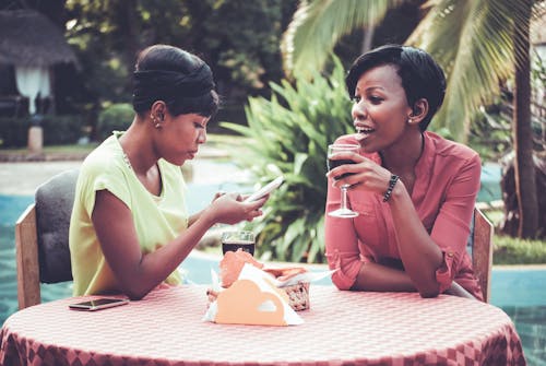 Woman Holding Wine Glass Talking To Woman Using Smartphone