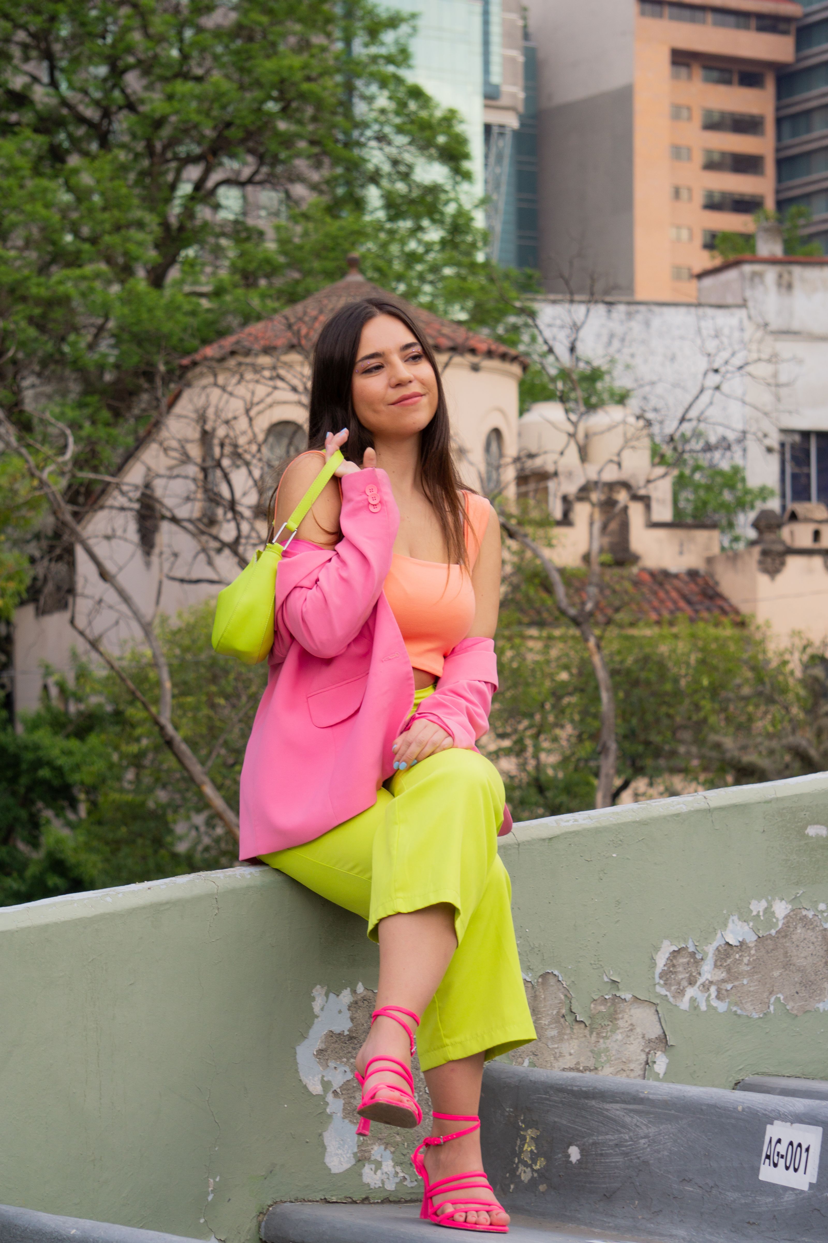 Mulher jovem e estilosa vestindo um traje neon sentada ao ar livre com um pano de fundo urbano, exalando estilo moderno.