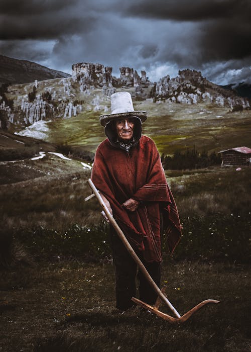 An Elderly Person in a Poncho and Hat Standing on a Grass Field under a Cloudy Sky 