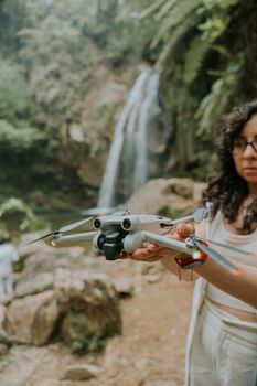 Woman holding a drone near a lush waterfall in a forest setting.