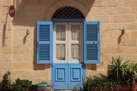 A picturesque blue shuttered window on a sunlit facade in Valletta, Malta, framed by potted plants.
