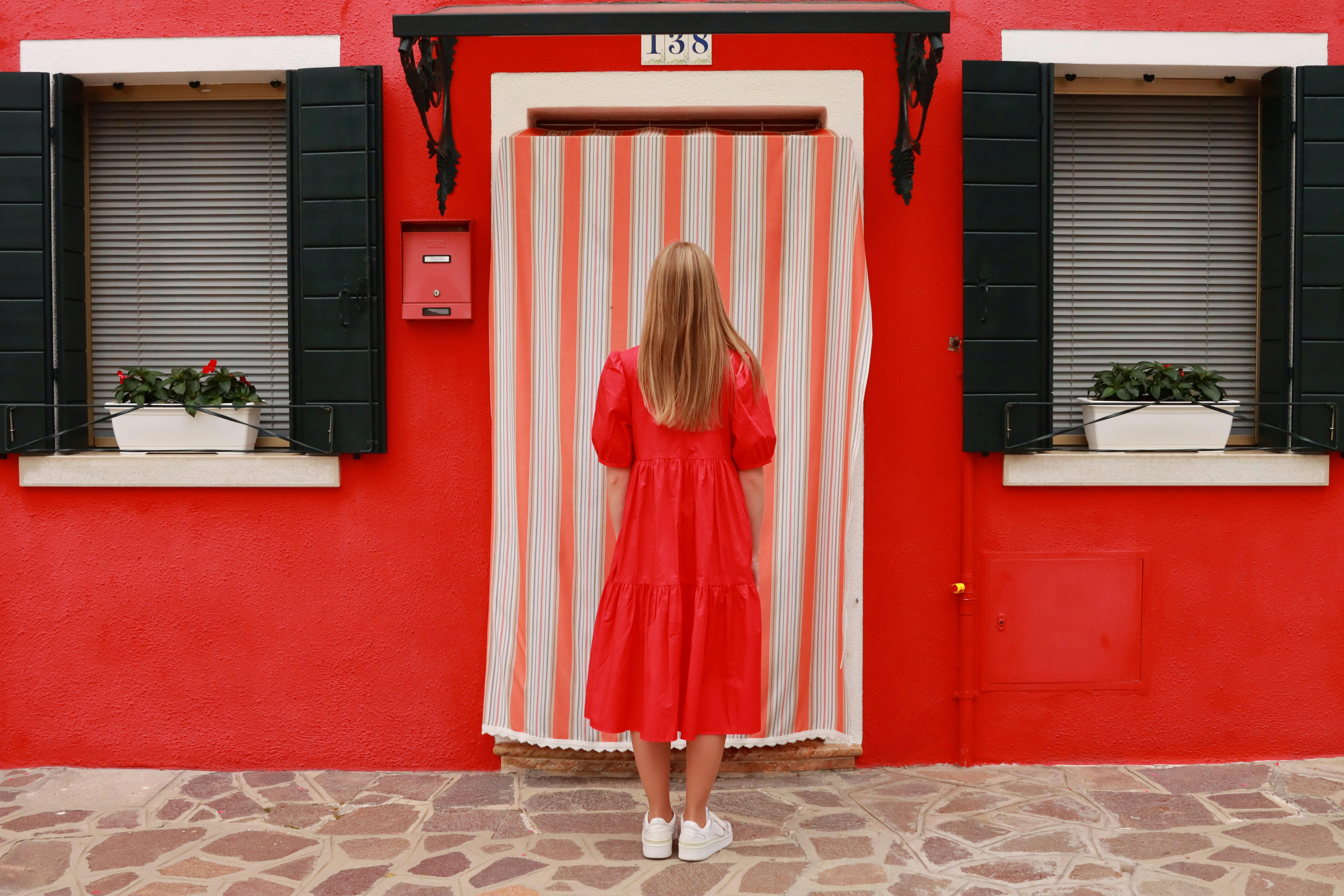 Woman in red dress standing in front of a vibrant red house with striped curtains in Venice, Italy.