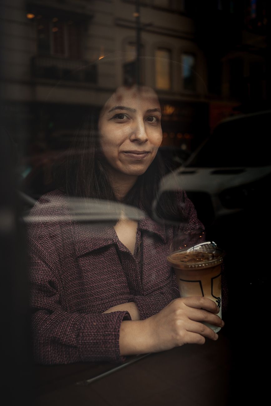 A woman relaxes with coffee by a window in a cozy urban café, captured through a reflective glass.