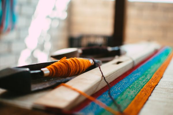 Weaving shuttle wound with orange yarn resting on a loom beam beside a striped textile — photo by Jorge Acre on Pexels