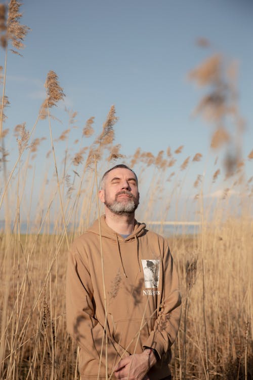 Man with a Gray Beard Wearing a Brown Hoodie Standing among Dry, High Grass