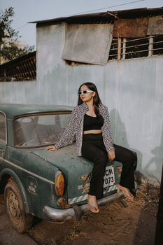 Young woman in retro fashion with sunglasses poses on an old car outdoors.