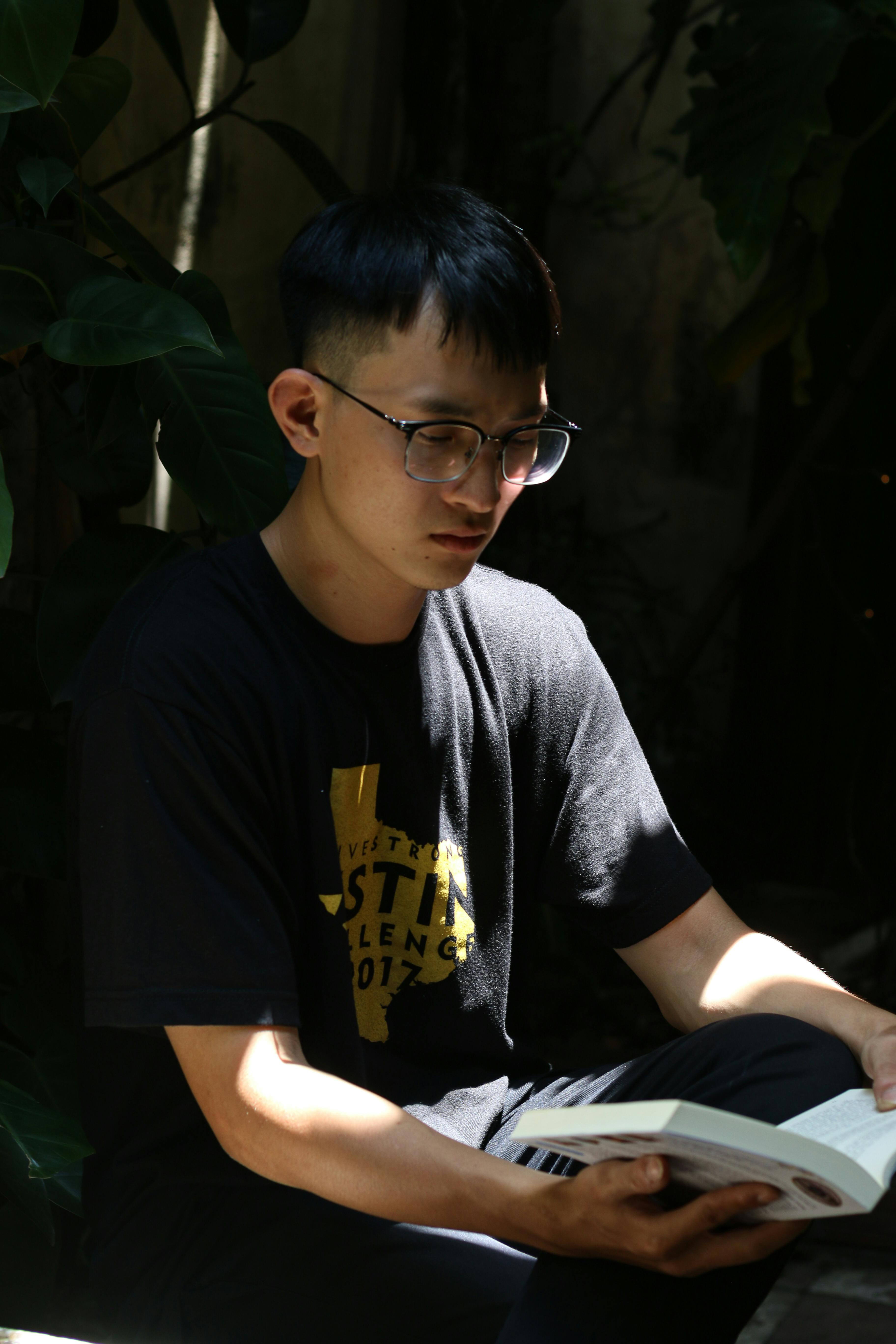A young man with glasses reads a book outdoors in dappled sunlight.