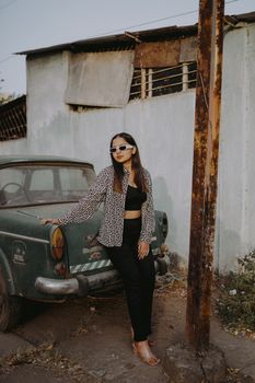 Young woman in retro outfit and sunglasses leaning on an old car, exuding vintage style.