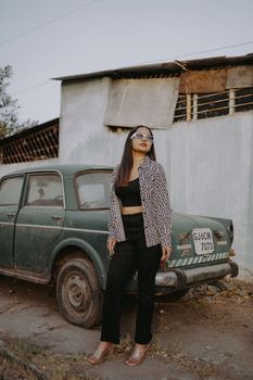 Stylish woman with sunglasses posing by a vintage car against a weathered wall.