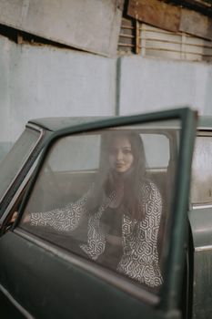 A woman in a patterned blouse poses inside a vintage car with an open door, displaying urban style.