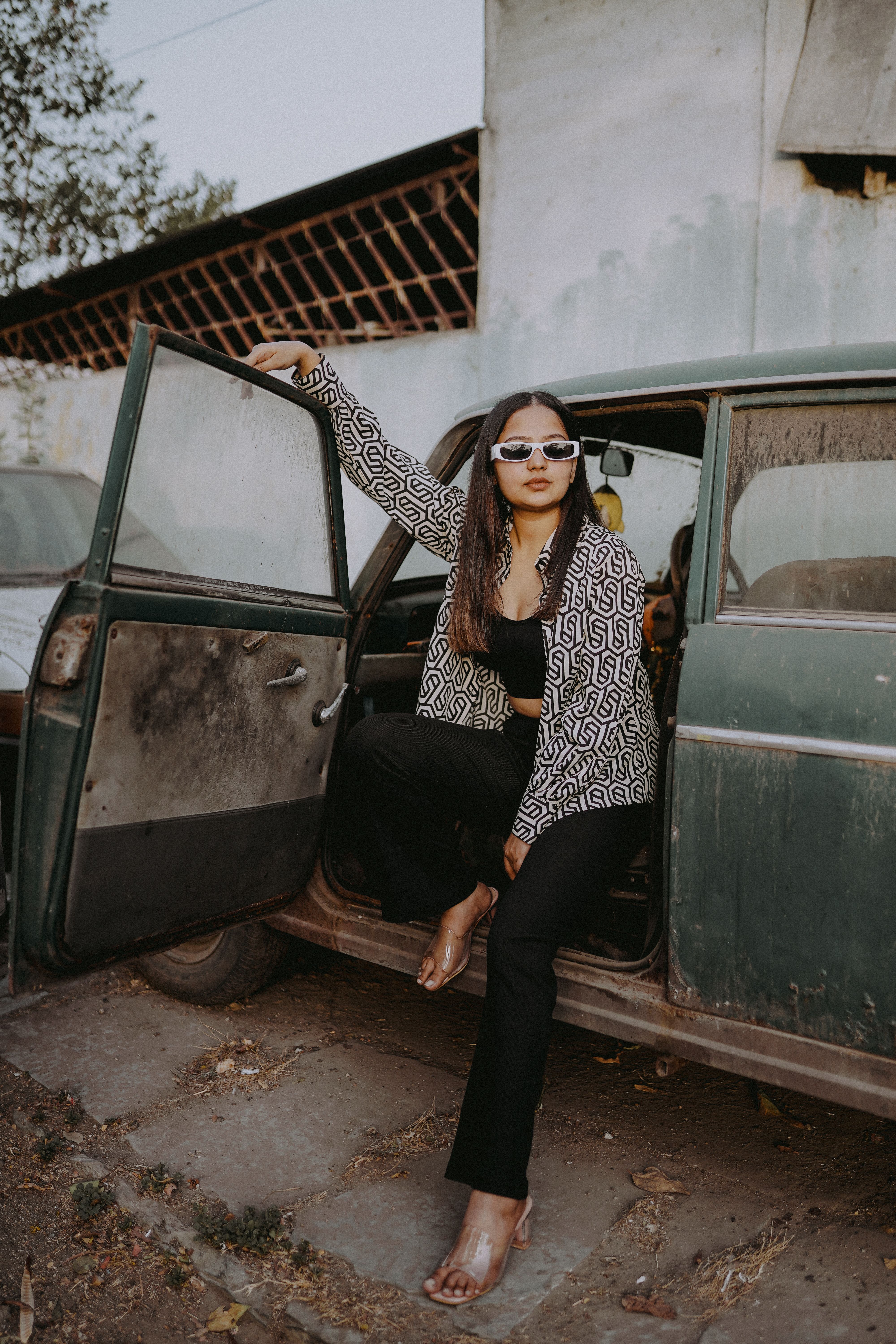 Fashionable woman in sunglasses posing by an old car, showcasing urban style outdoors.