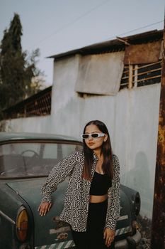 Fashionable young woman in sunglasses leaning on a vintage car outdoors in urban area.