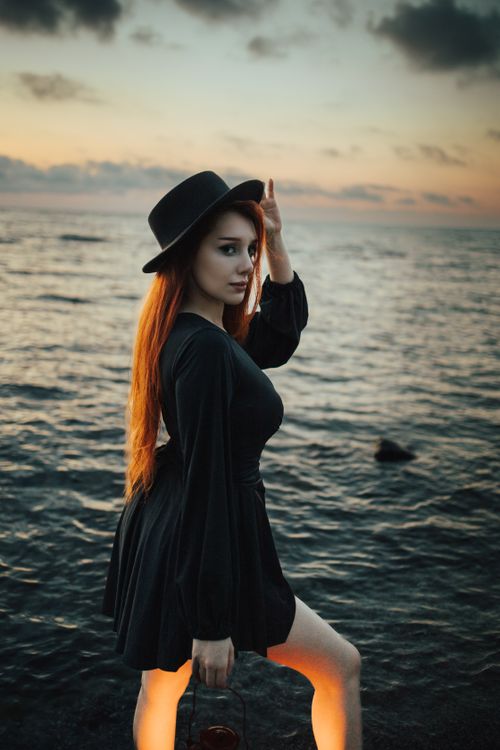 Free A stylish woman in a black dress poses by the sea at sunset, exuding modern elegance. Stock Photo