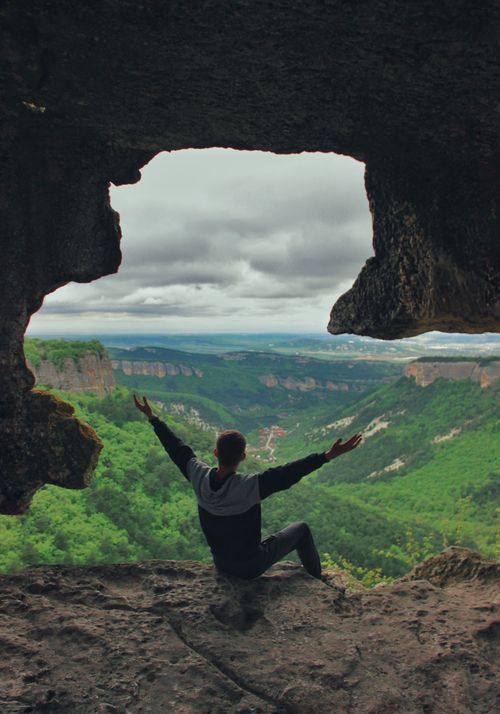 Free Man sitting with arms raised inside a cave, overlooking a lush, expansive valley. Stock Photo