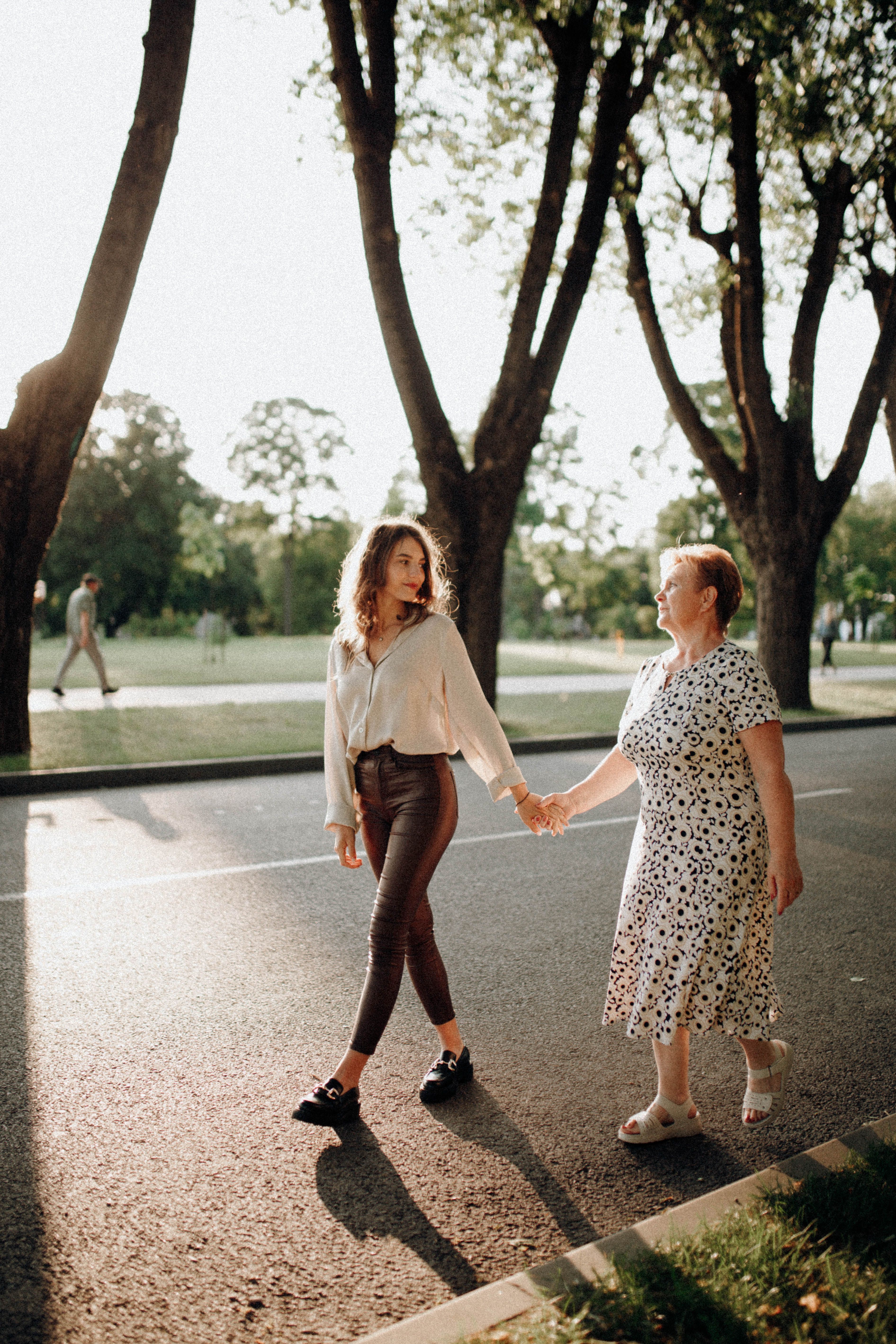 A grandmother and granddaughter walk hand in hand down a sunny park pathway, enjoying a summer day.