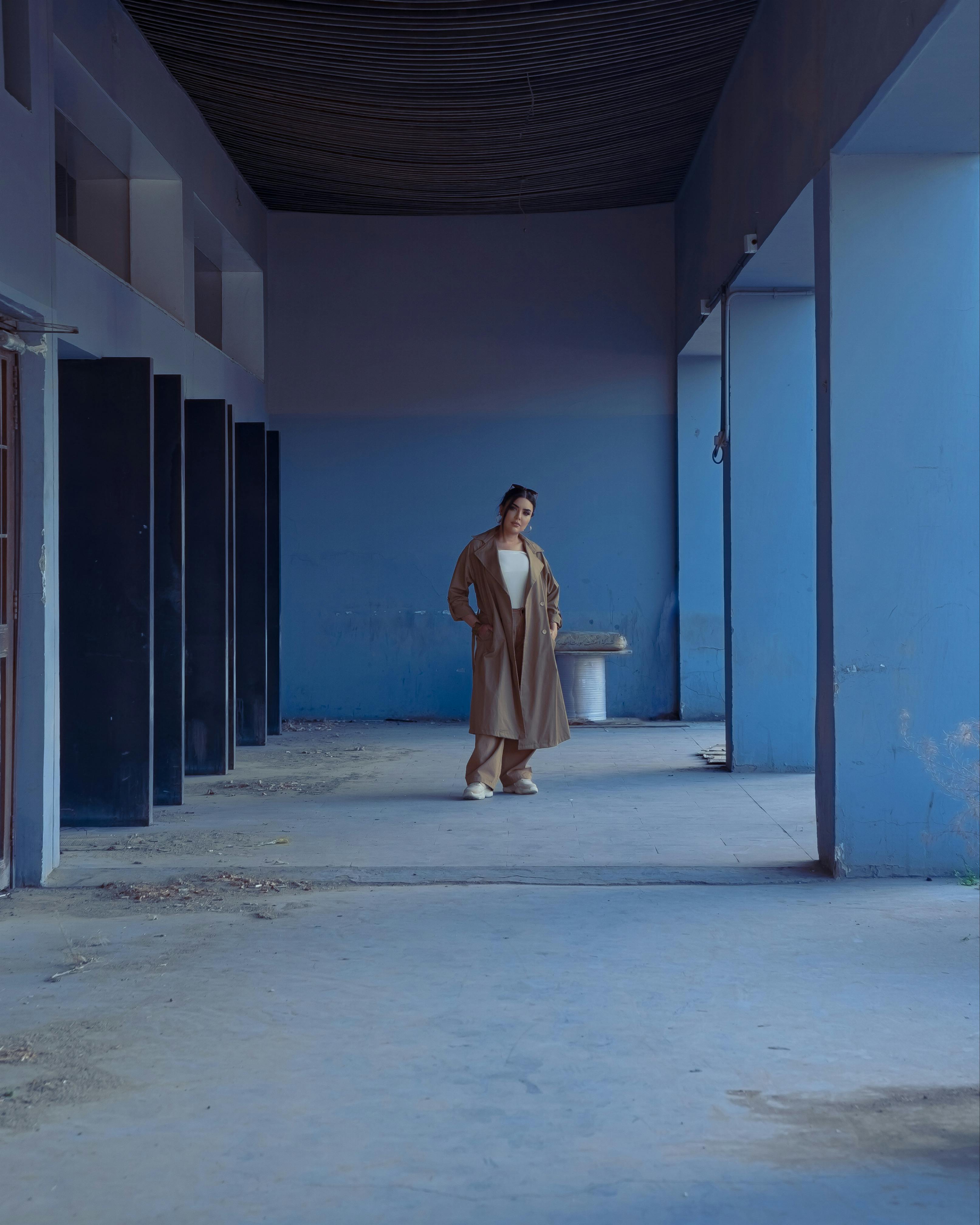 Fashionable woman in coat poses in urban corridor with blue walls, creating a modern and dramatic scene.