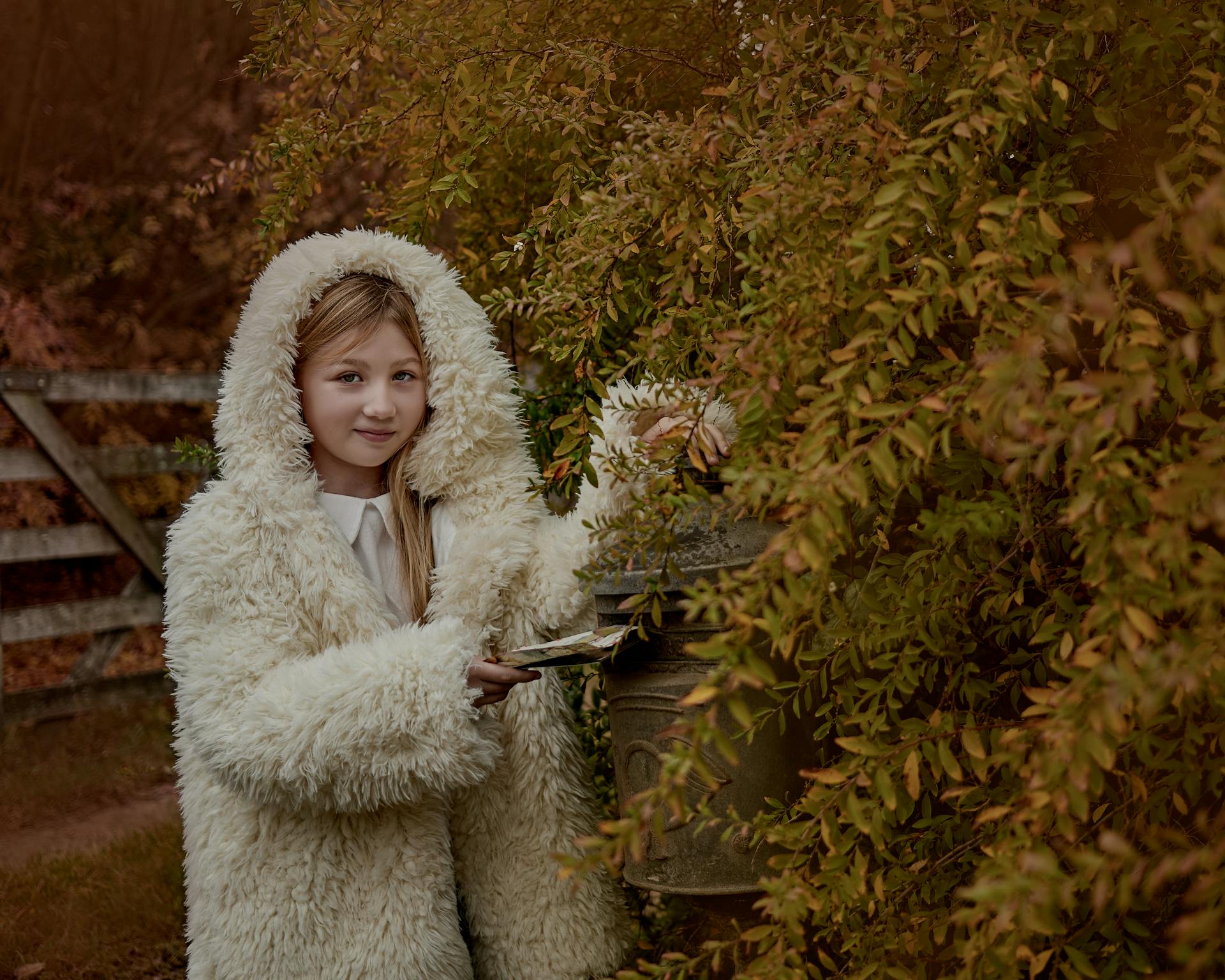 A girl wearing a fluffy fur coat smiles as she posts mail outdoors in a leafy setting.