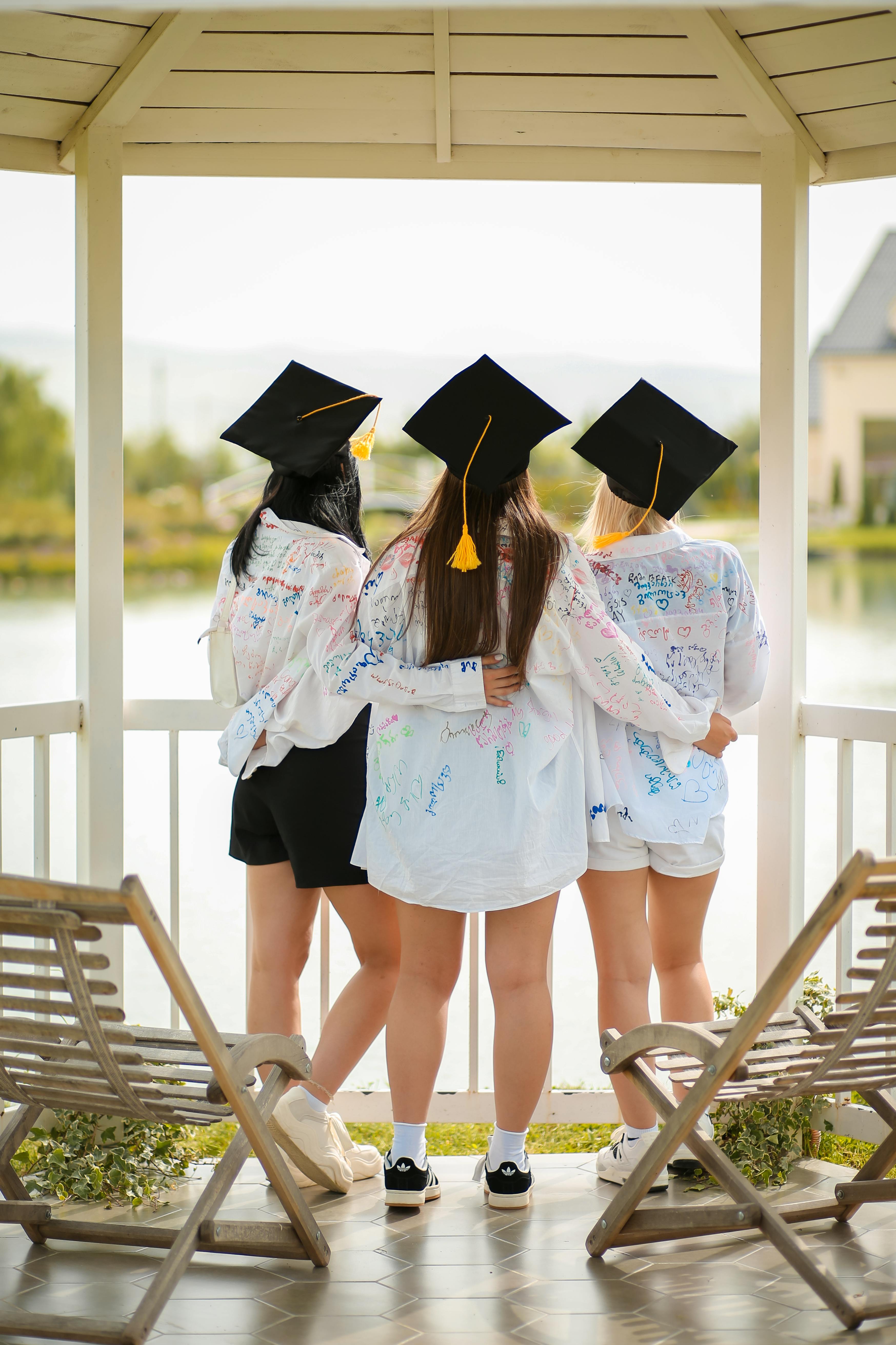 Three female graduates in mortarboards embrace, celebrating outdoors in a gazebo by a lake.