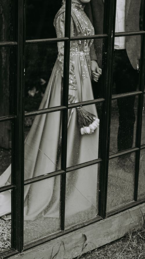 Free Elegant couple holding hands by a window, dressed in formal attire, captured in black and white. Stock Photo