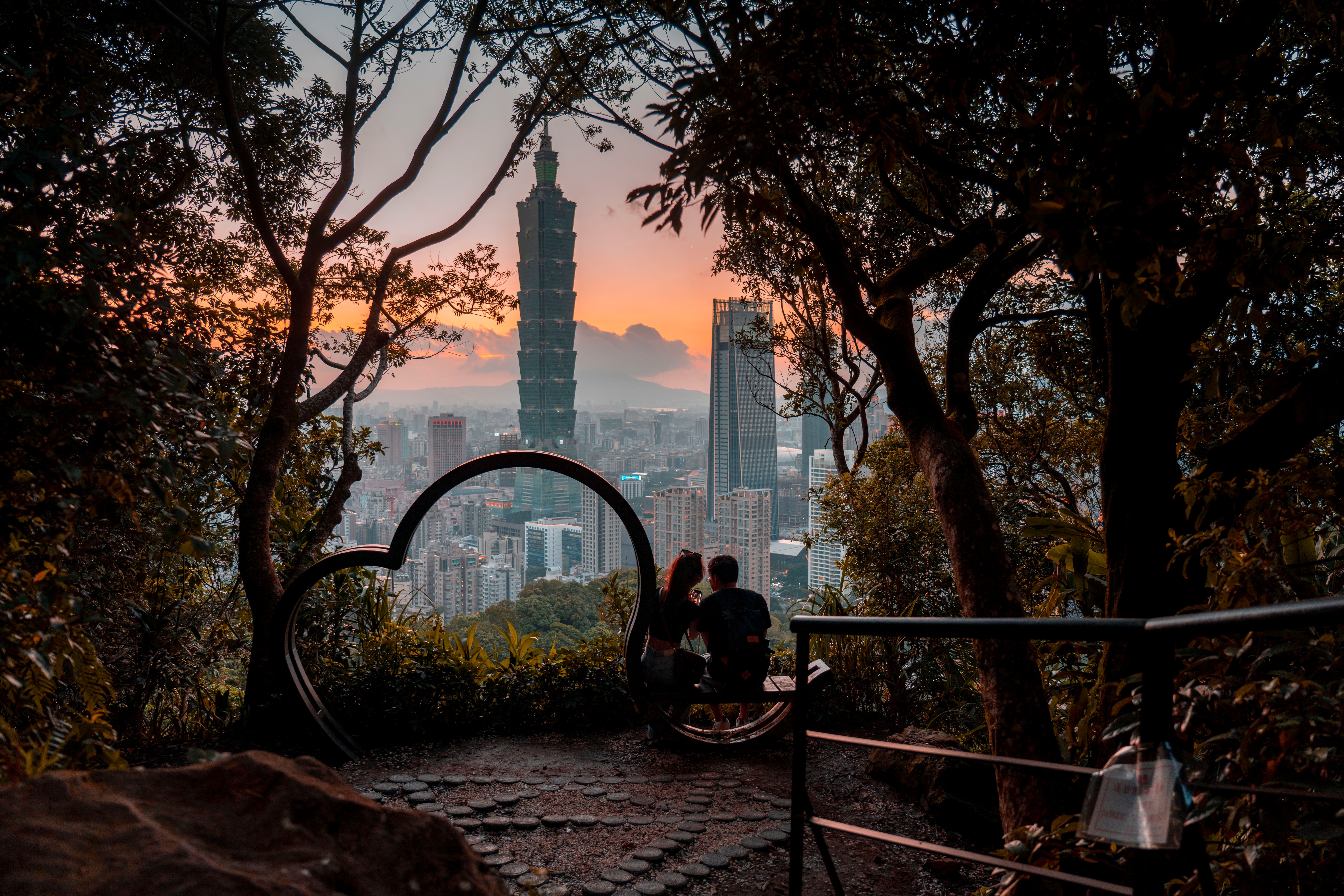 A couple sits on a scenic hill, admiring Taipei 101 at sunset, framed by lush trees.