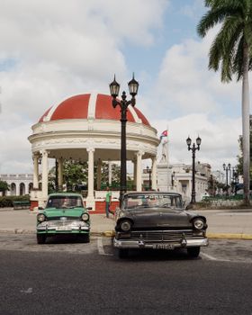 Classic Cuban scene with vintage cars and a historic pavilion in a sunny city square.