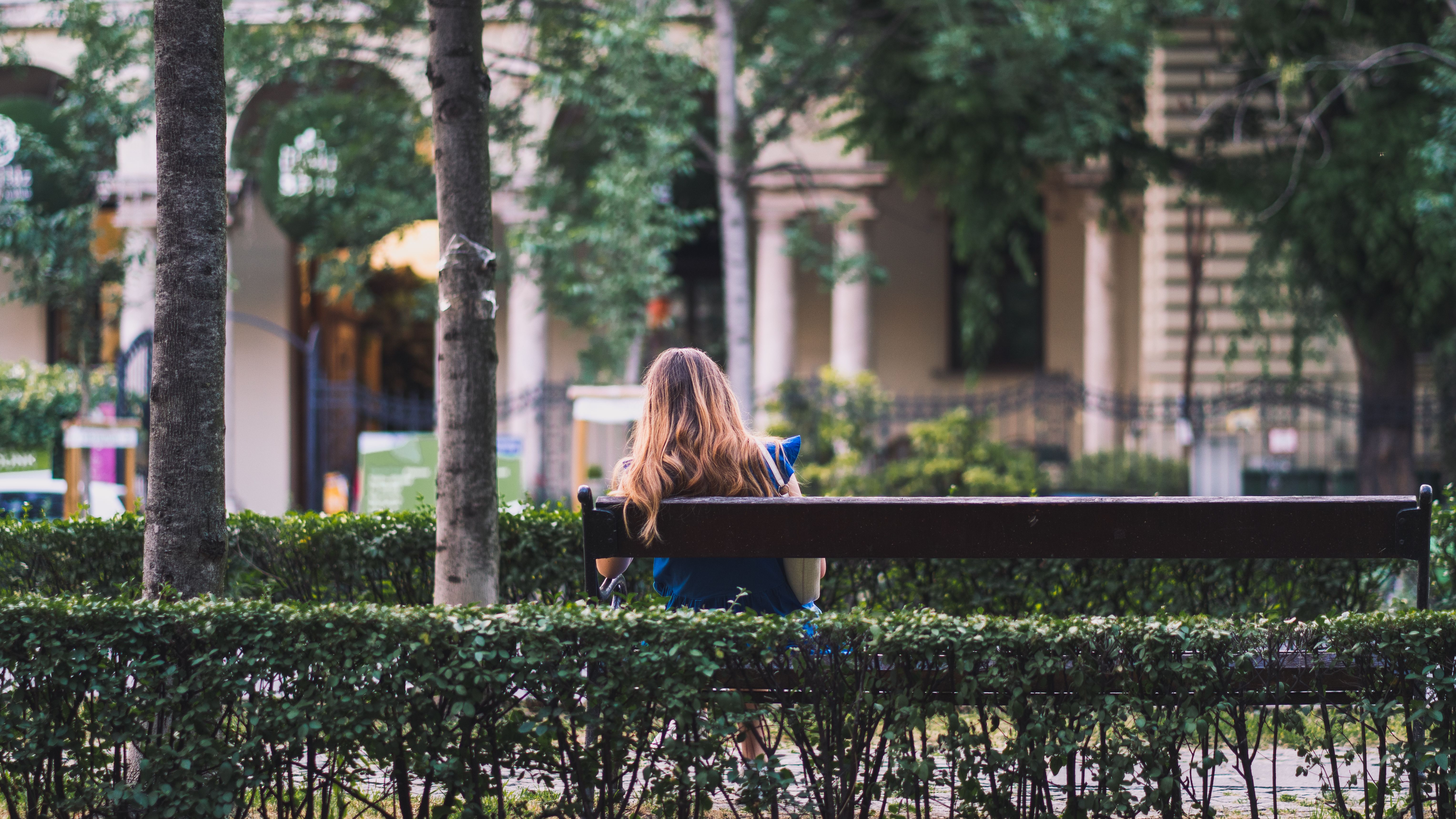 A woman with long hair reads a book while sitting on a bench in an urban park setting.