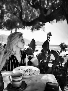 一位优雅的女士戴着墨镜，穿着别致的服装，在海边咖啡馆享用美食，享受轻松的生活方式