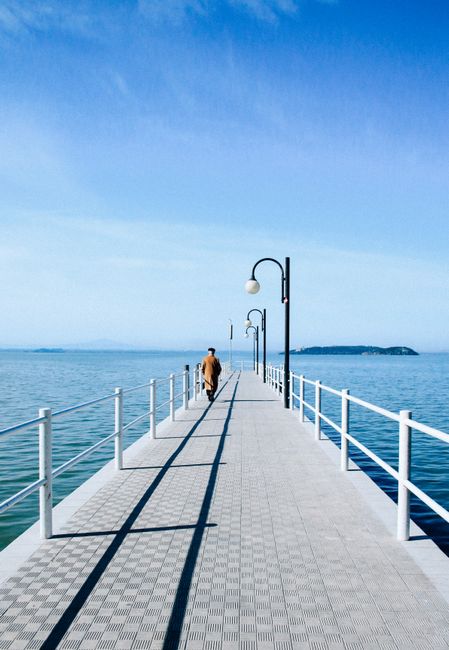 A lone figure walks along a pier extending into the tranquil sea under a vivid blue sky.