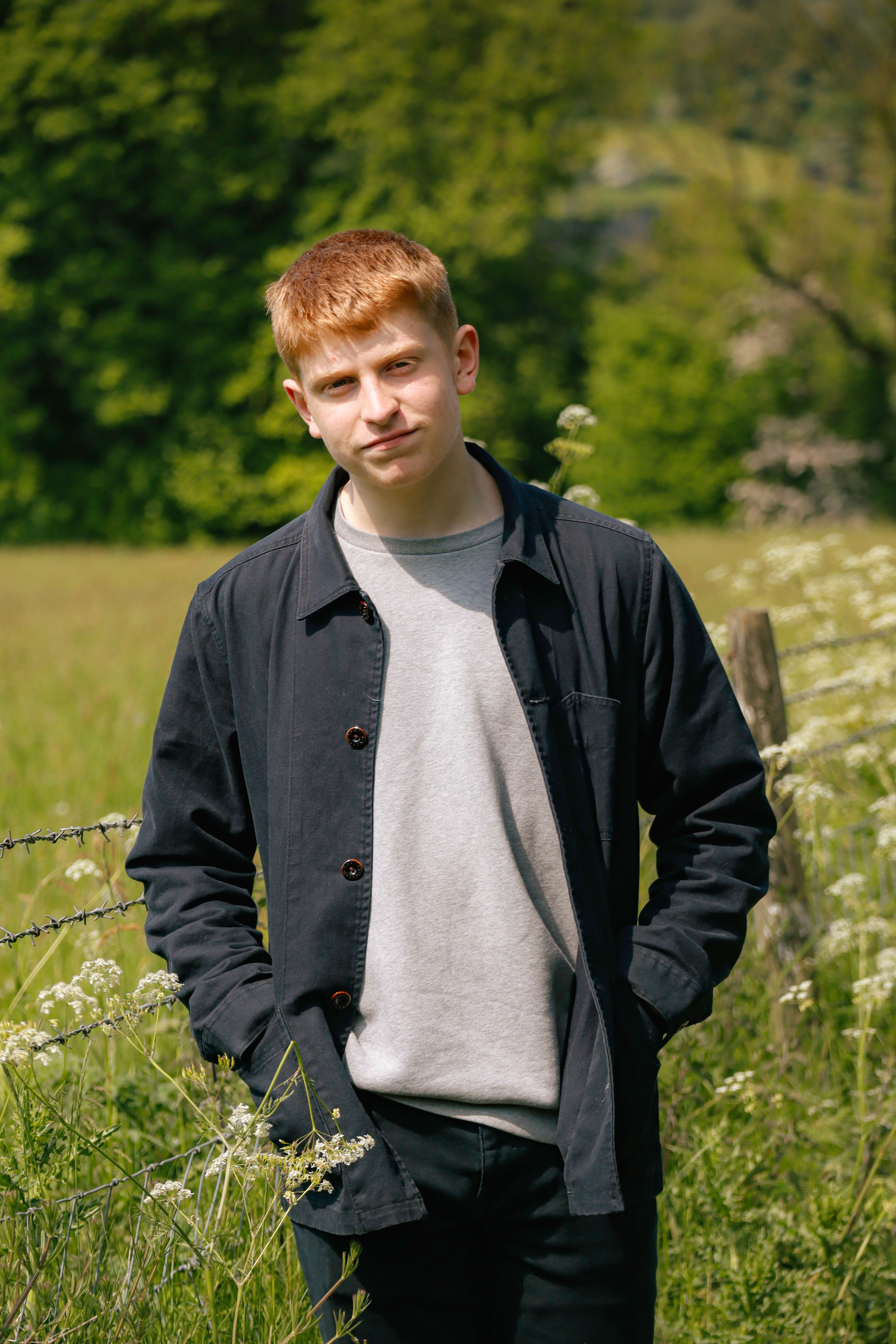 A young man in a casual outfit poses in a lush green spring meadow with flowers and a fence.