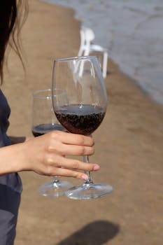 A close-up of hands holding wine glasses on a sandy beach with chairs in the background.