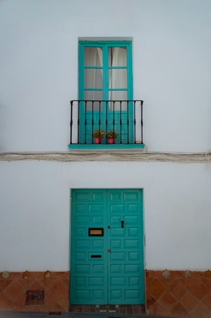 Minimalist facade featuring turquoise doors and balcony against a white wall, accented with potted plants.