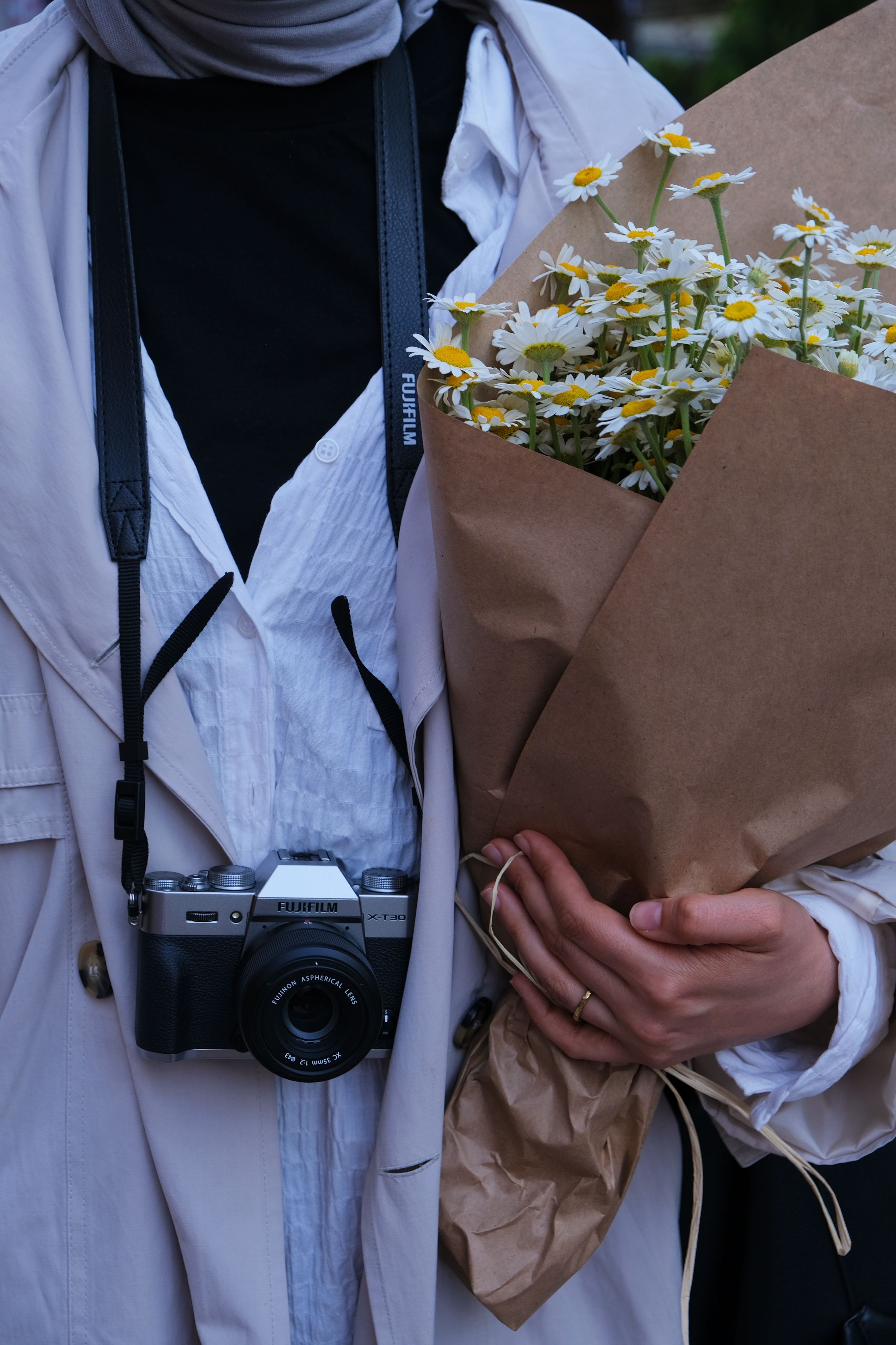 Chic woman in light coat holds a vintage camera and bouquet of daisies, embracing nature and photography.