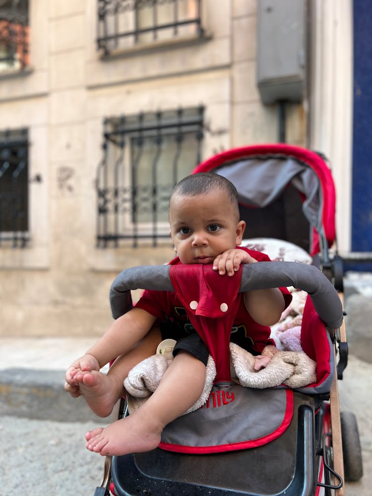 Small Baby Boy Sitting In A Red And Gray Stroller On A Street