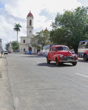 A classic red car driving on a city street in Cuba with colonial architecture.