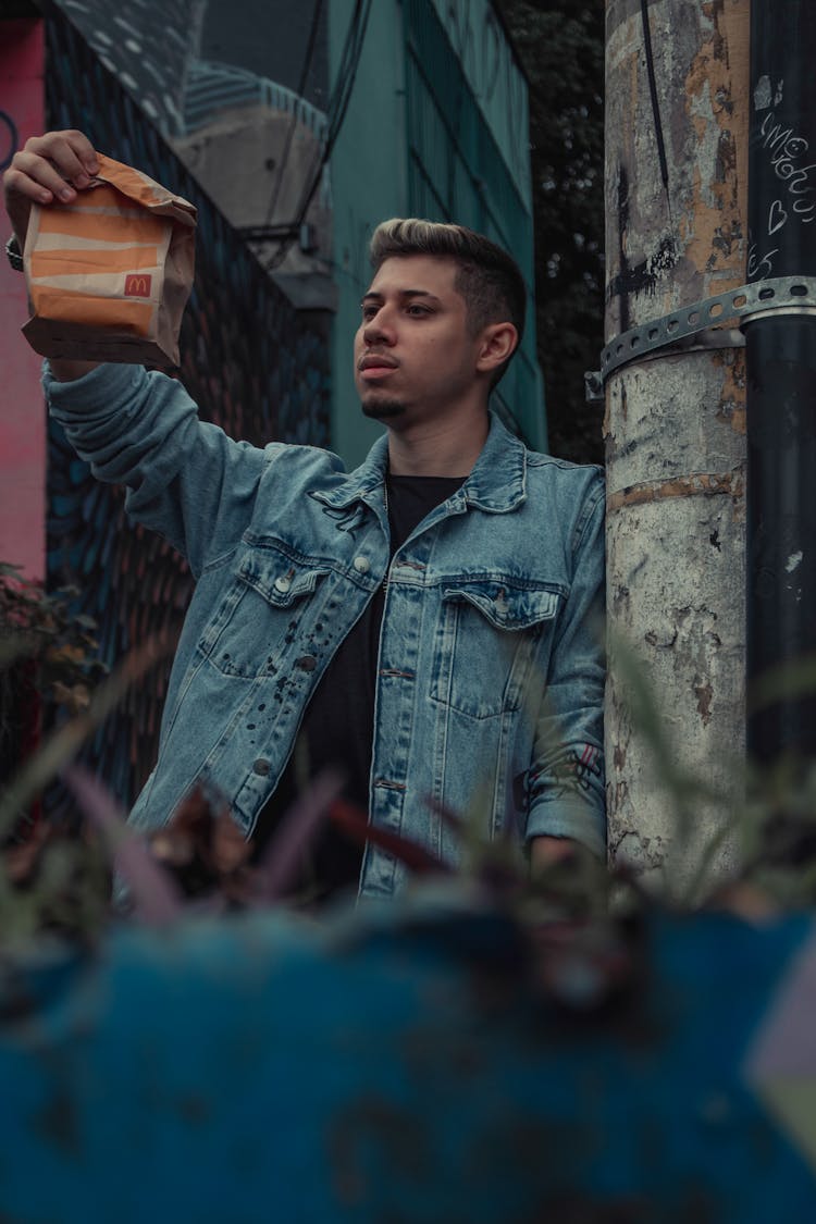 Young Man In A Denim Jacket Holding A Paper Bag With Fast Food Takeaway 