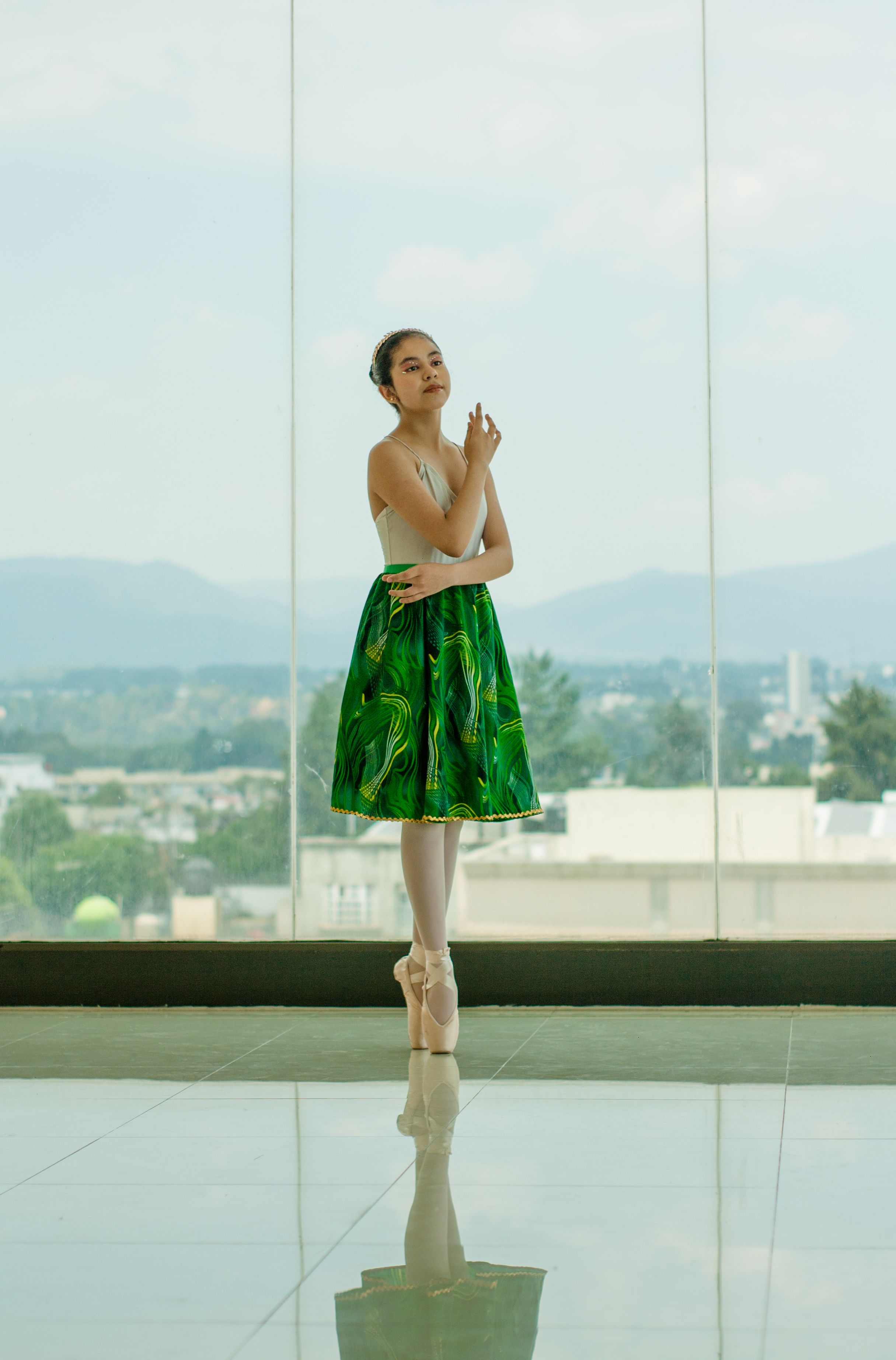 Elegant ballerina in green dress posing indoors by a large window with a scenic view.