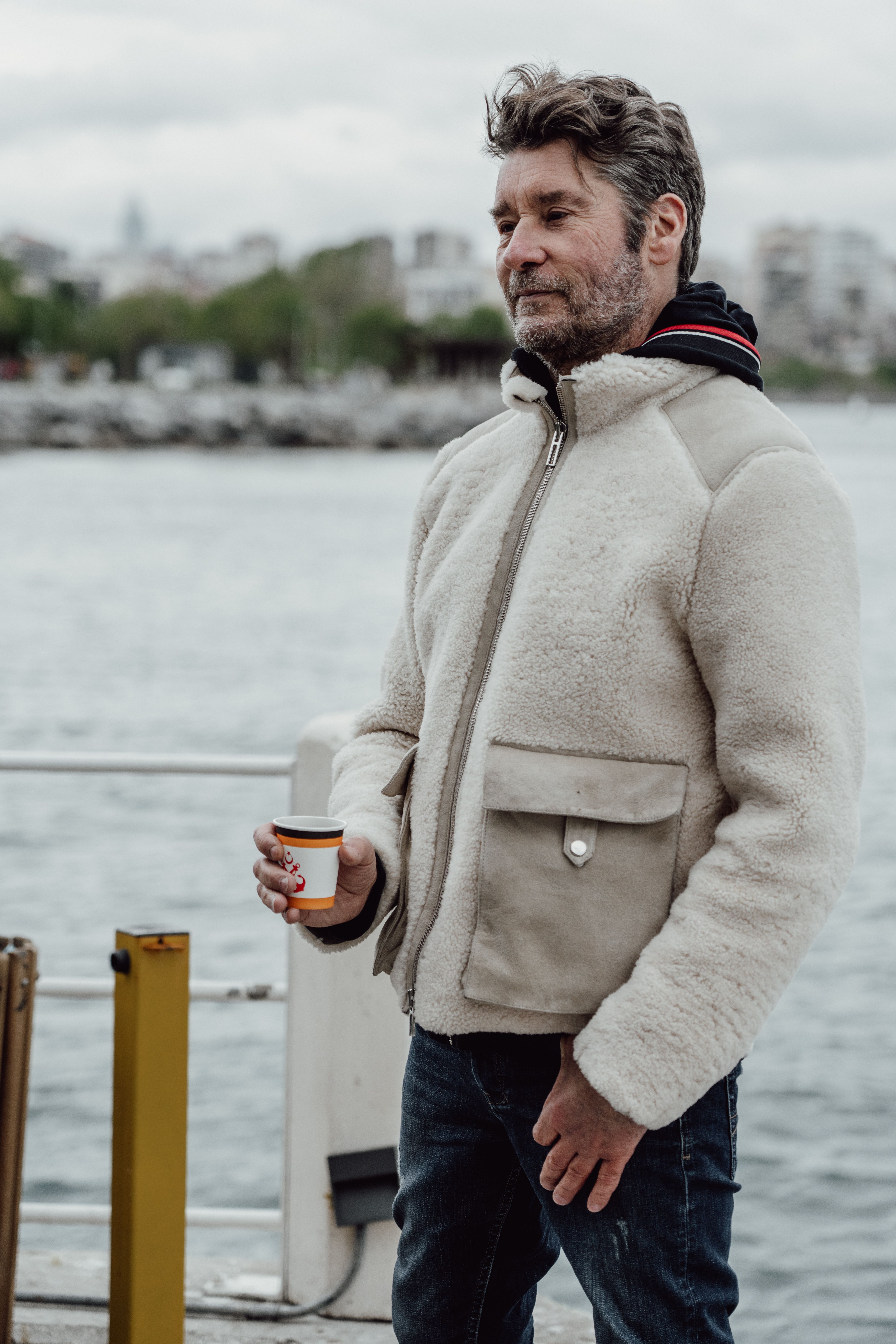 Adult man enjoys coffee outdoors near the sea, wearing a warm jacket.