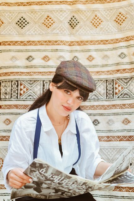 Woman in white shirt and cap reads newspaper against ornate tapestry backdrop, vintage style.