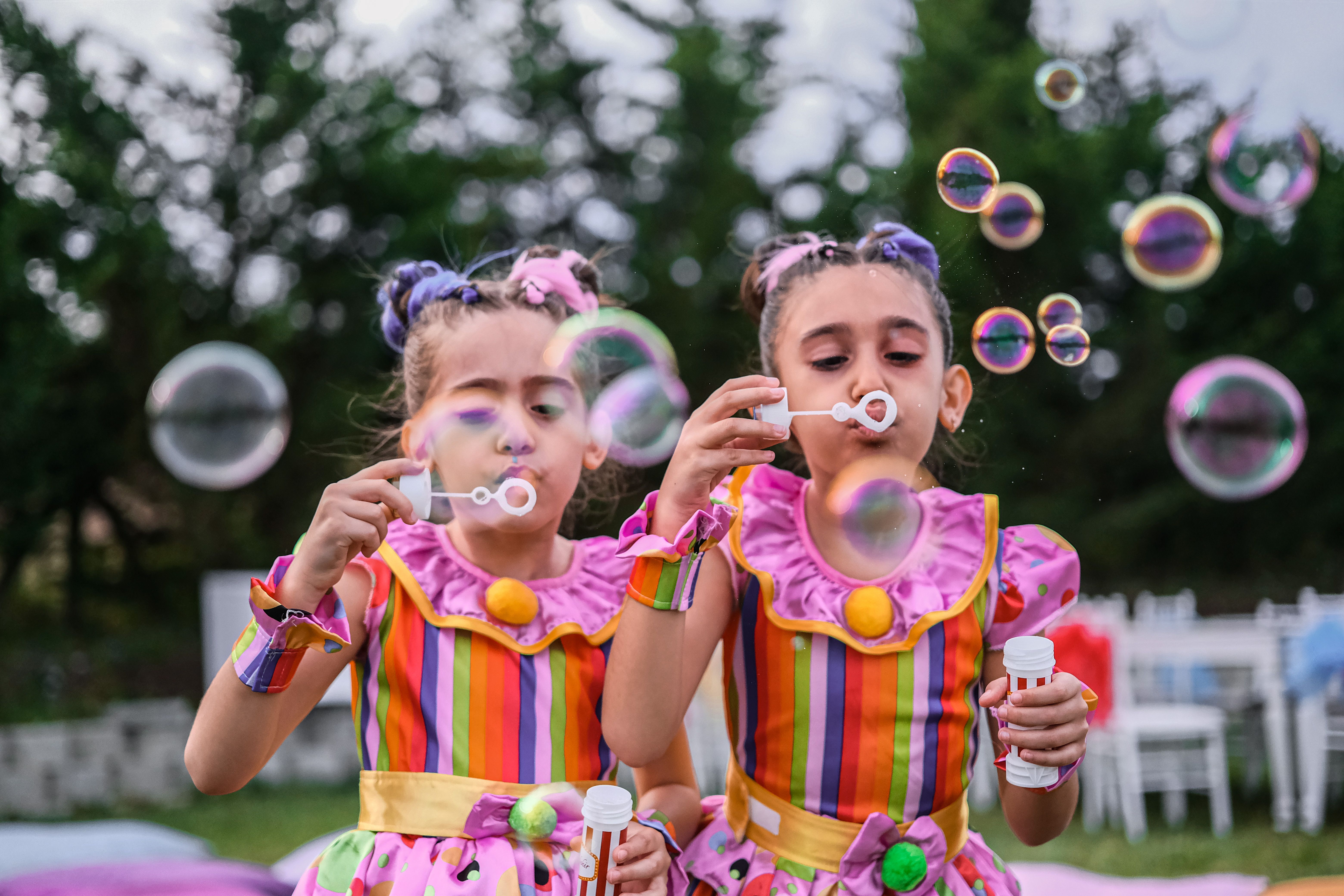 AUE event entertainment — Two young girls in colorful clothing enjoy blowing soap bubbles in a playful outdoor setting.