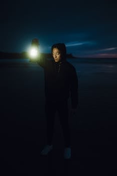 A young man in a hoodie holds an illuminated lantern on a dark beach at night.