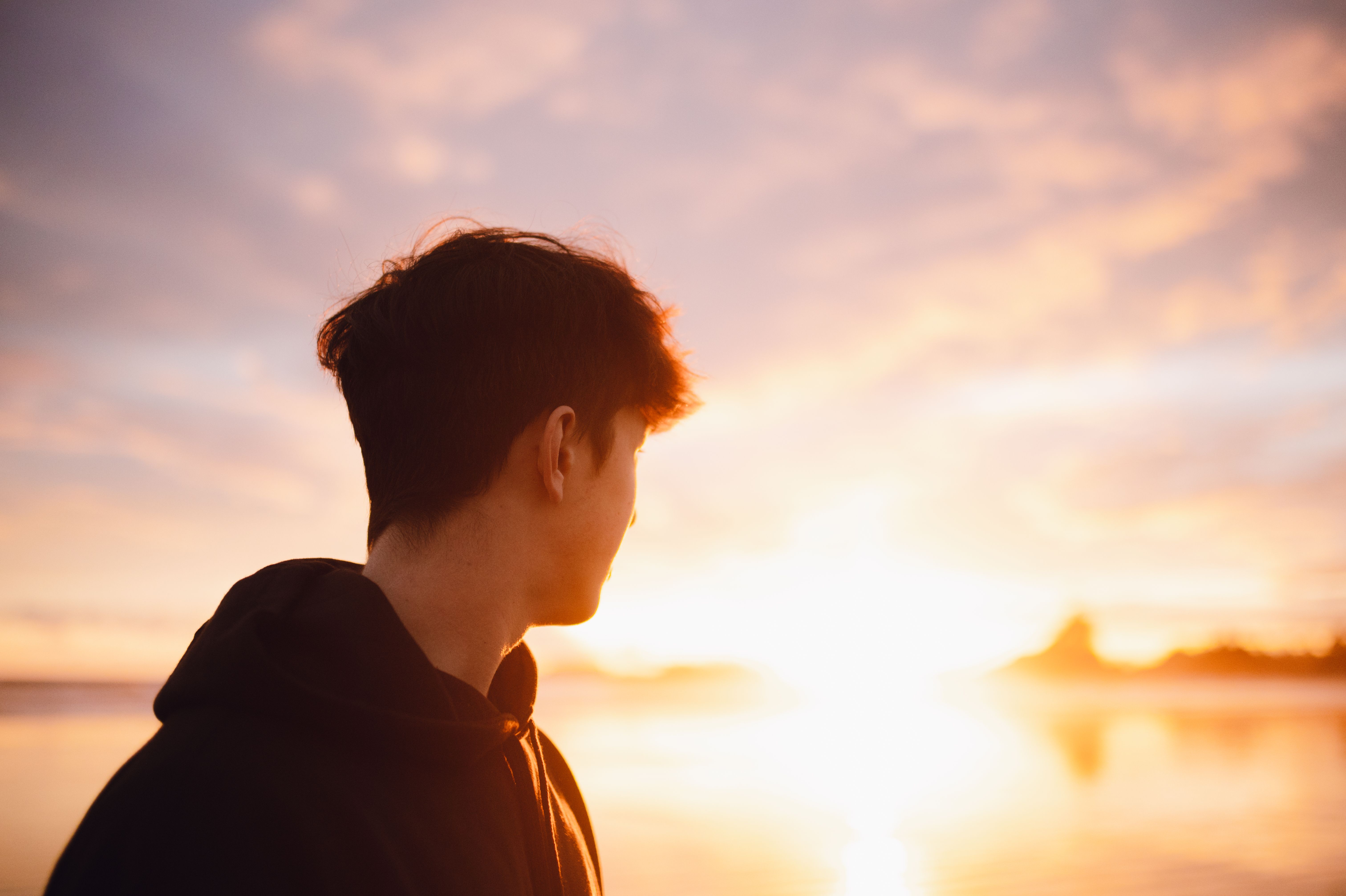 Silhouette of a young man in a hoodie looking at a scenic sunset by the beach.