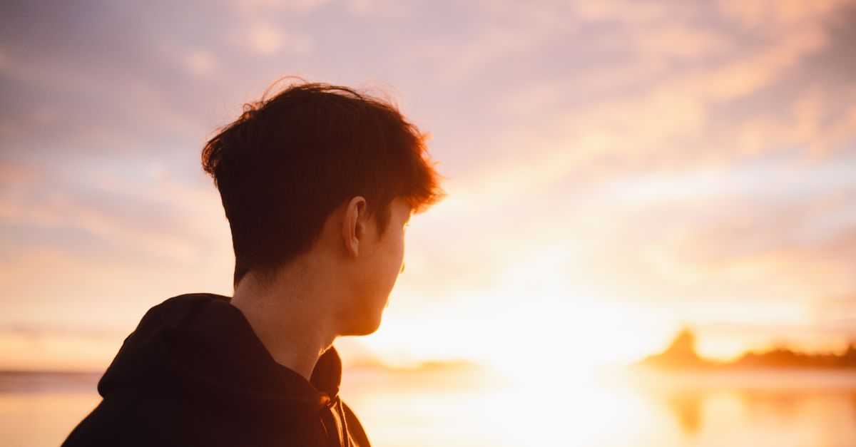 Silhouette of a young man in a hoodie looking at a scenic sunset by the beach.